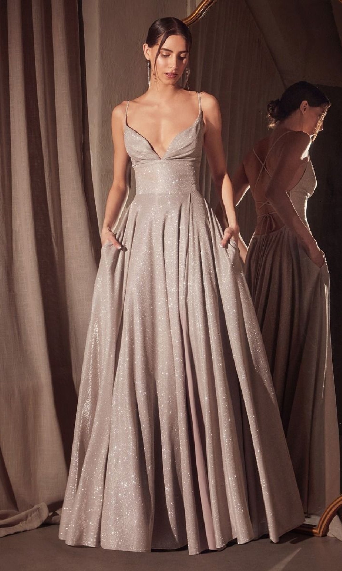 A woman in the Strappy-Back Glitter Prom Gown: Amoris T214, featuring shimmering silver fabric, thin straps, a deep neckline, and dress pockets, stands before a mirror with her hands in the pockets.